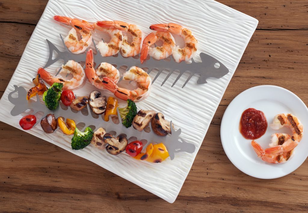 Skewers of grilled shrimp and veggies sit on a serving platter, skewered y the stainless steel slide & serve bbq skewers from Proud Grill company