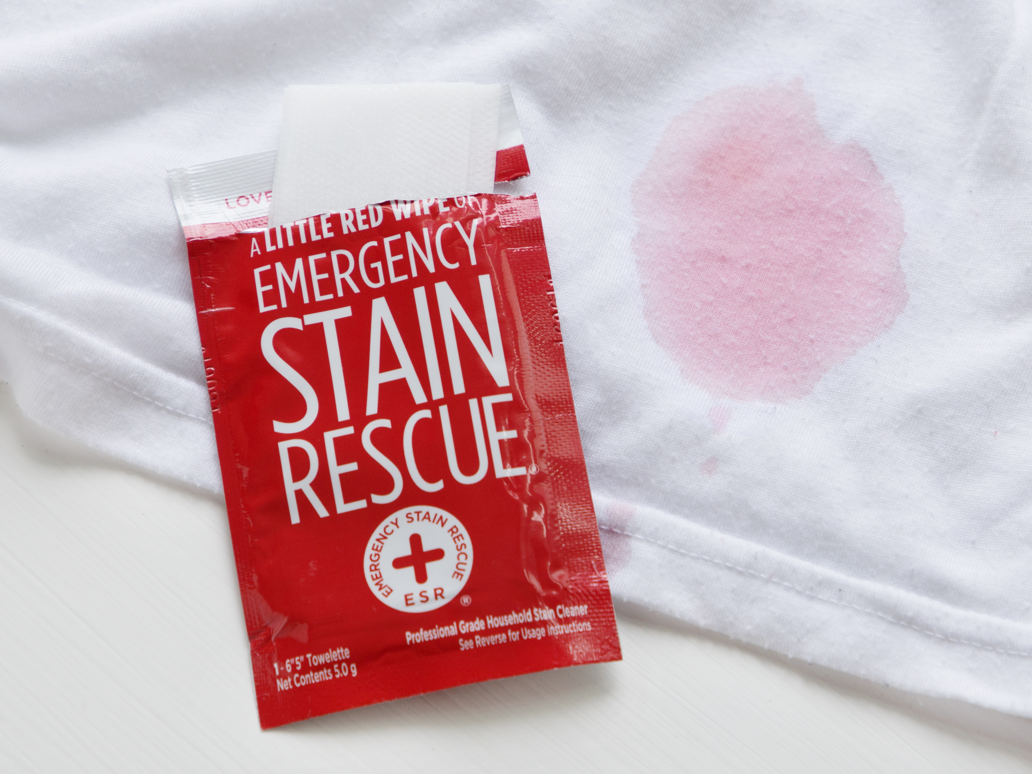 A little red wipe of emergency stain rescue sits next to a white shirt stained with red wine