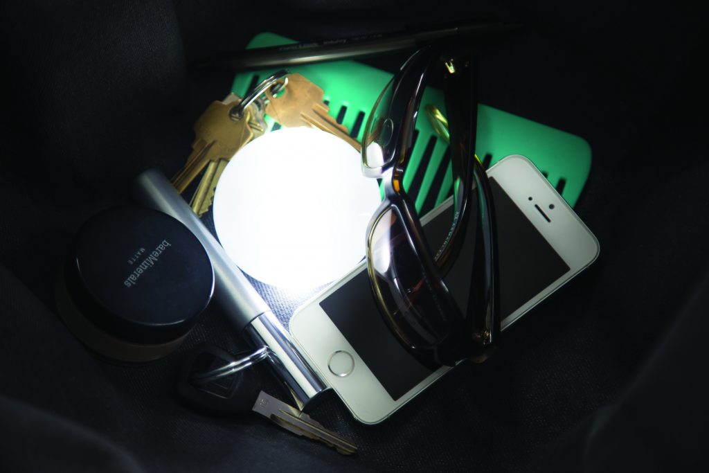 A SOI automatic handbag light sits lit in the bottom of a purse surrounded by keys, makeup, glasses & a phone