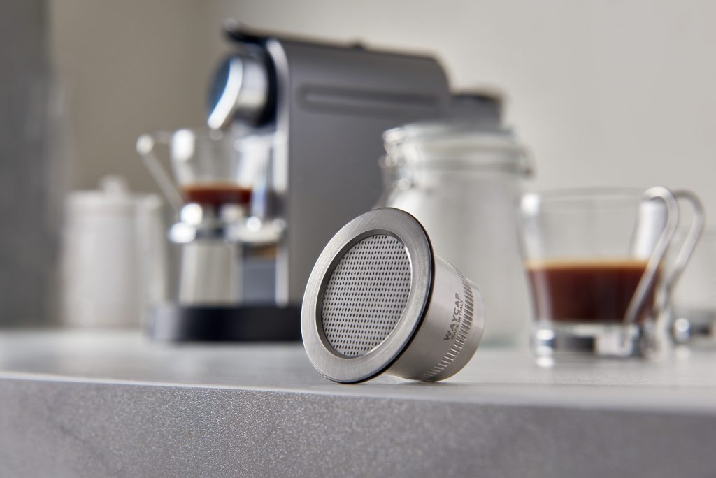 A Waycup refillable Nespresso capsule resting on a kitchen counter in front of a Nespresso machine. 
