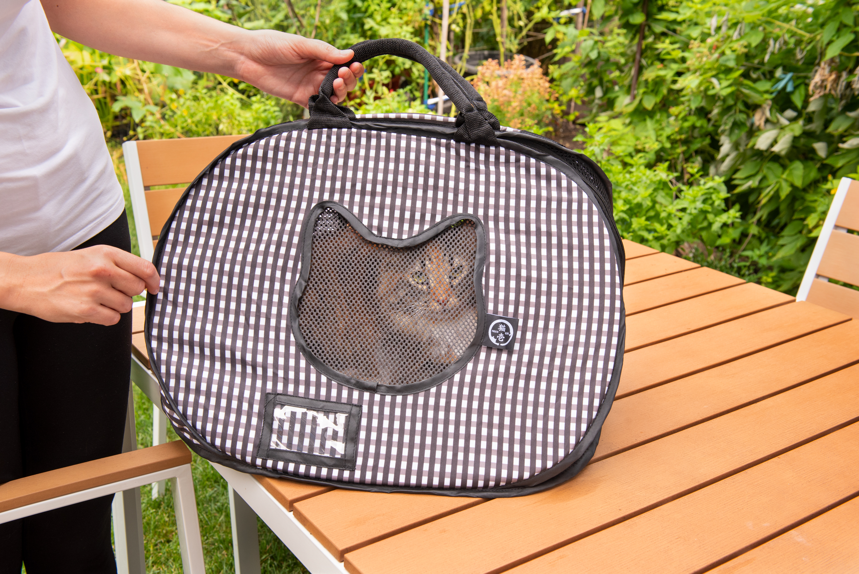 A Calico cat is seen sitting in a black & white checkered ultra-light collapsible cat carrier from Necoichi