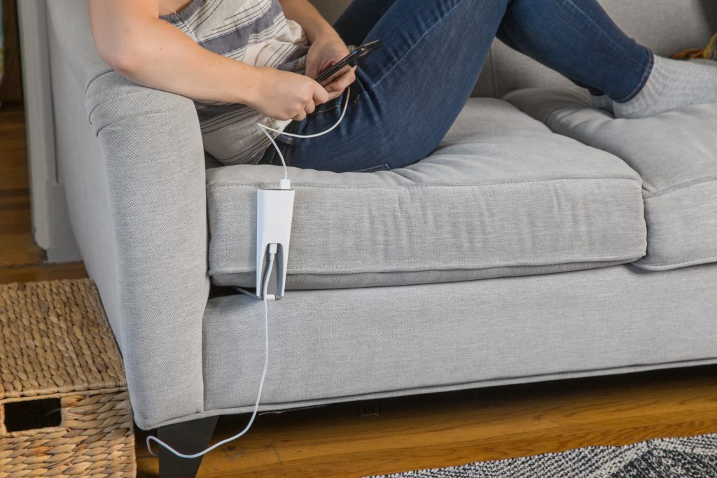 A person is seen relaxing on a couch, using a Couchlet couch phone charger to charge their phone