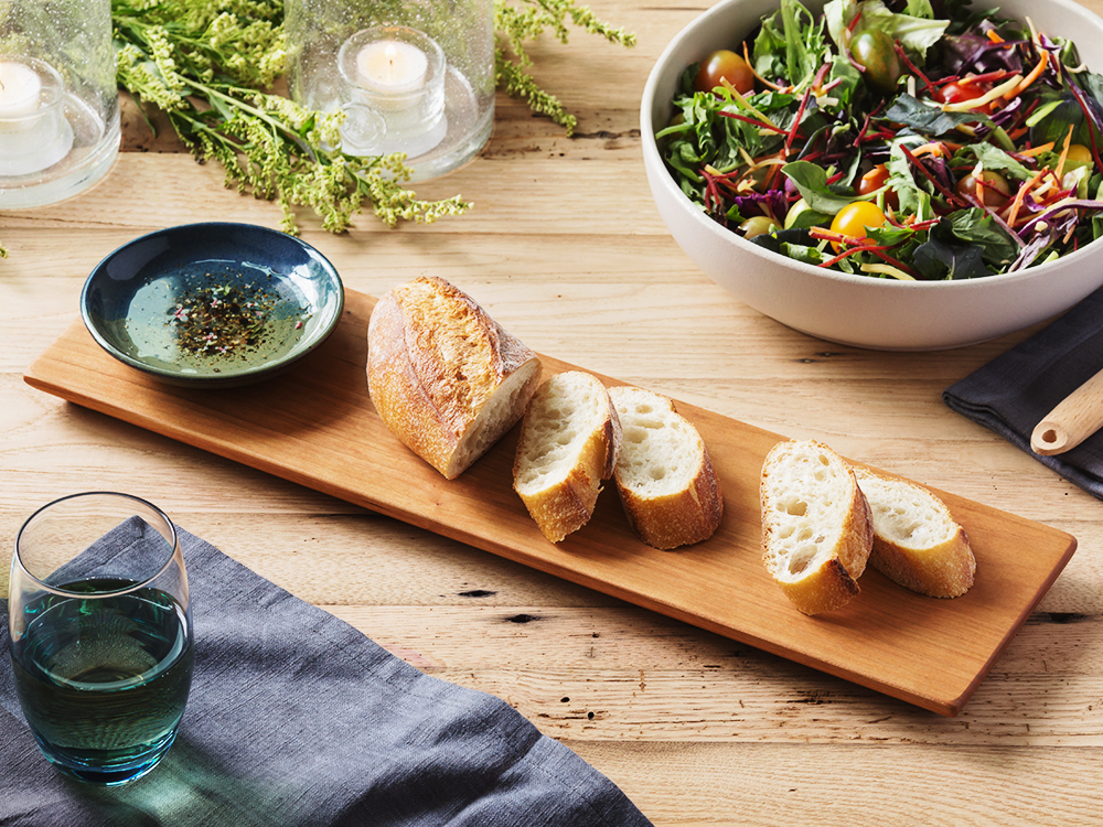 A wooden board with sliced pieces of bread, a ceramic dish with olive oil in it, and a salad sit on a counter.
