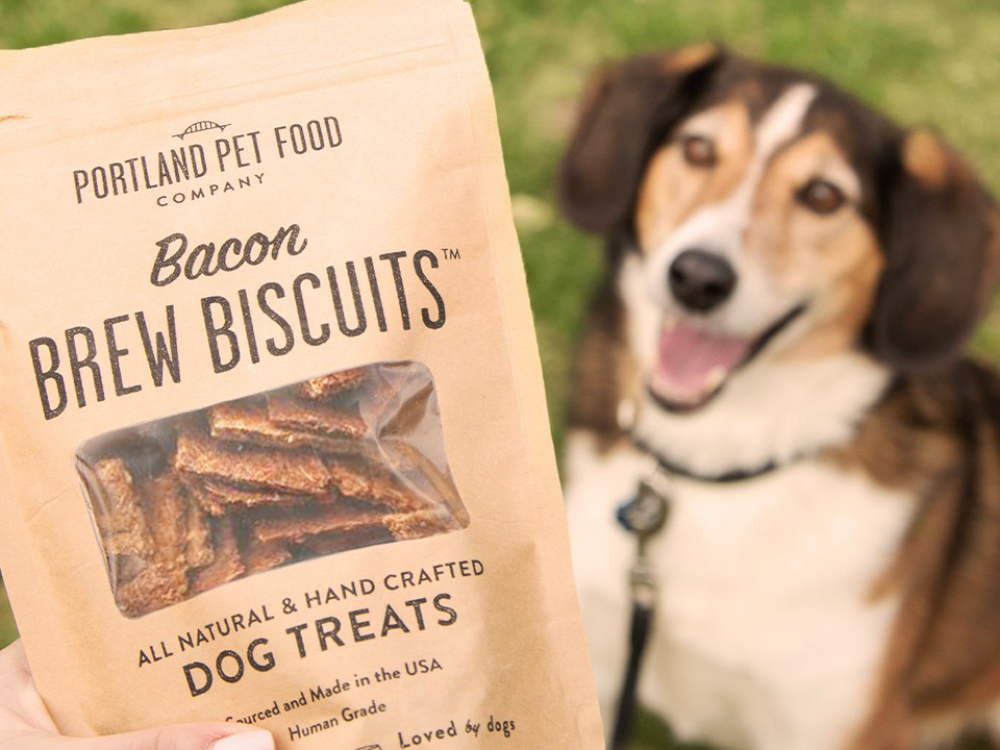 A dog is seen eagerly awaiting a bacon brew biscuit dog treat from Portland Pet Food Company