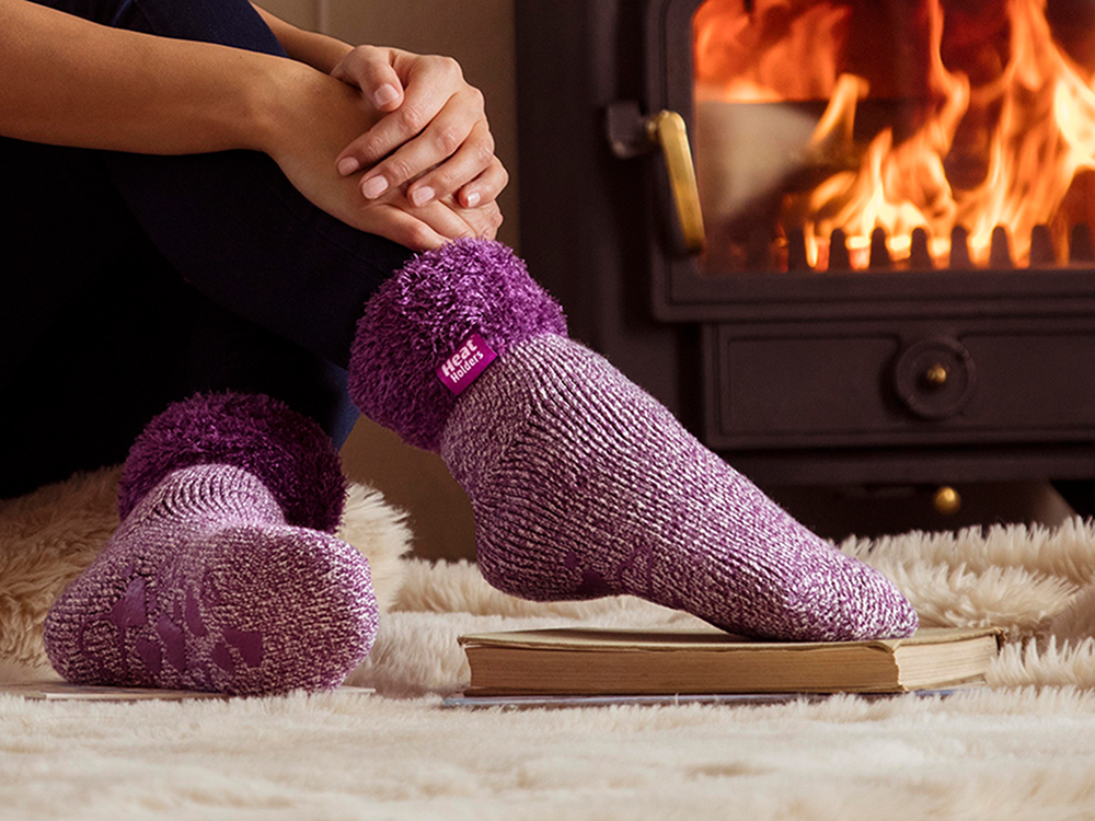 A woman is seen lounging by the fire wearing pink brushed thermal lounge socks from Heat Holders