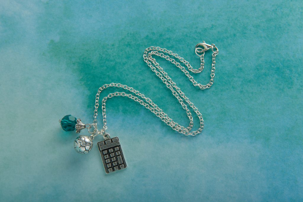 A Smart Girls Jewelry charm necklace features charms for soccer and a calculator with a teal bead