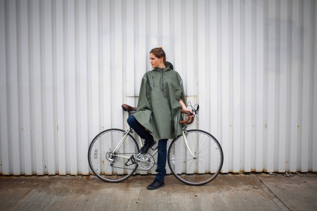 A woman is seen standing next to her bike wearing an olive green Cleverhood high-performance rain cape
